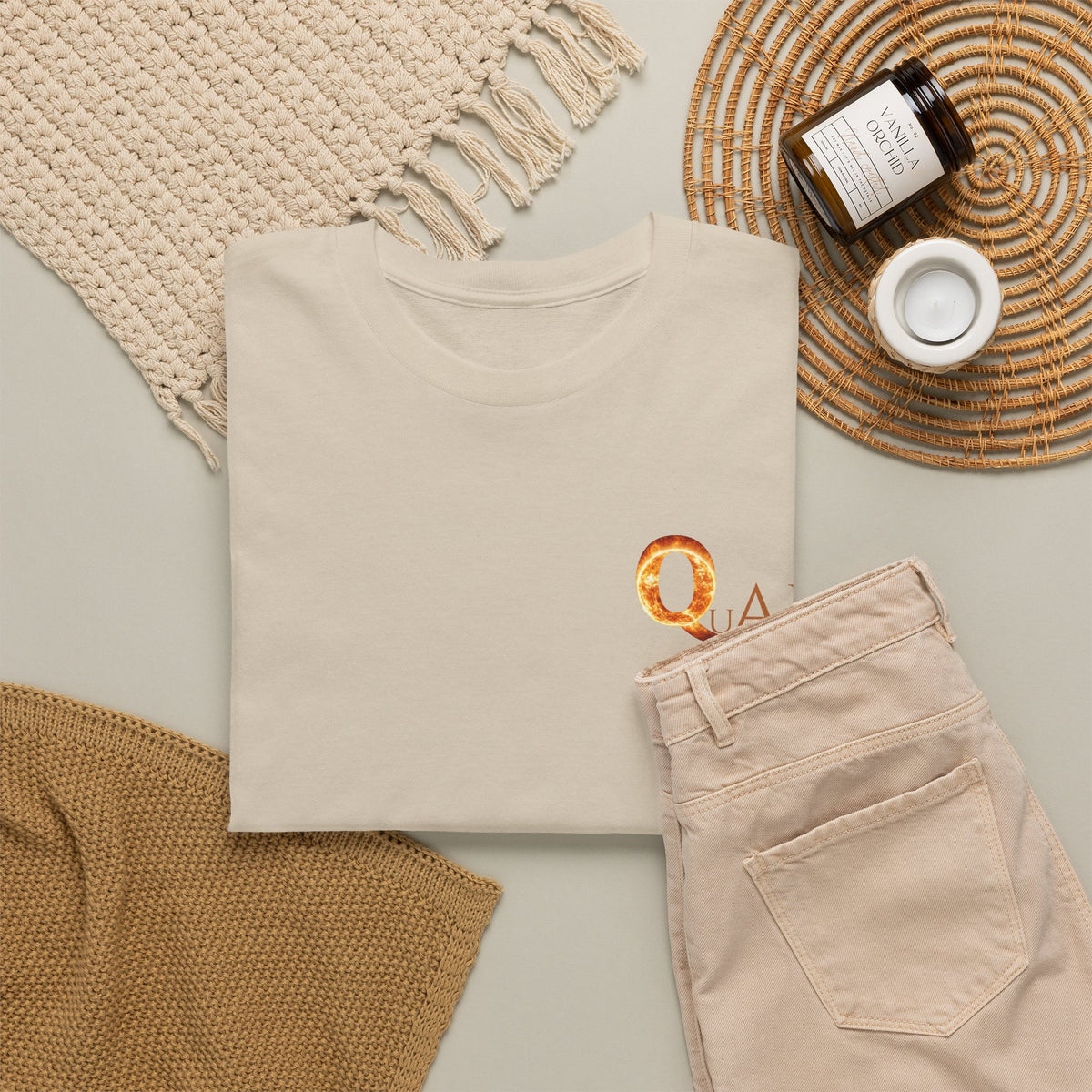A folded cream-colored t-shirt with a small QuAH graphic on the chest, styled in a flat lay with beige pants, a woven blanket, a candle jar, and a small candle on a neutral background.